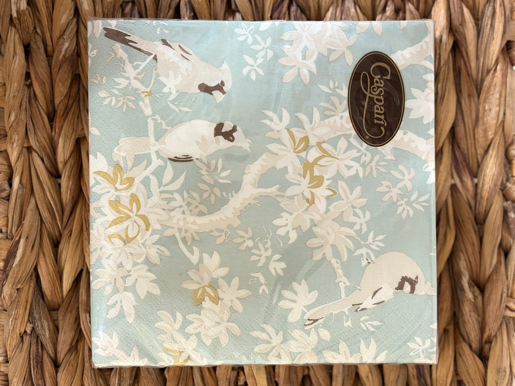 Servilleta extendida con aves posadas entre hojas blancas y doradas sobre fondo celeste