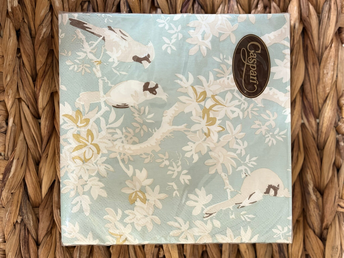 Servilleta extendida con aves posadas entre hojas blancas y doradas sobre fondo celeste